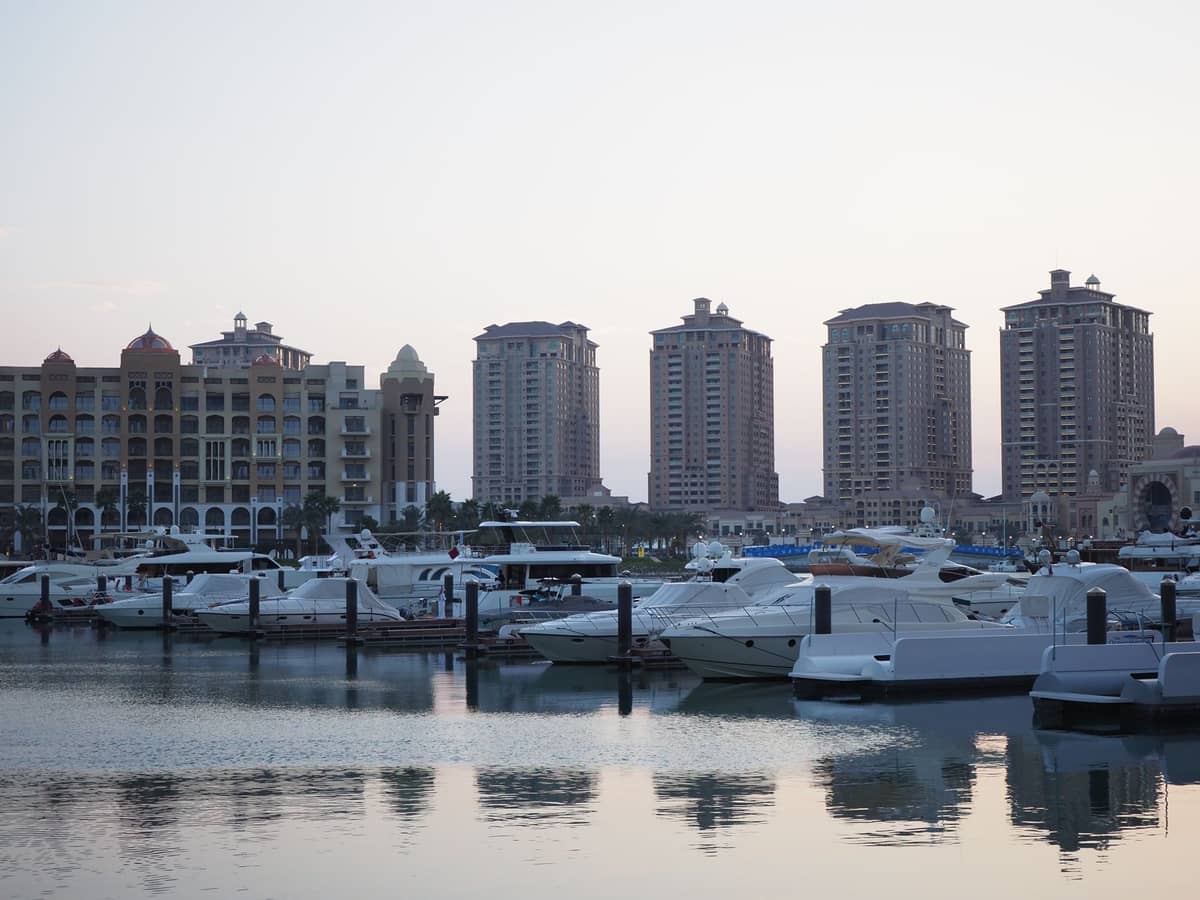 Exklusive Marina mit Luxusyachten entlang der Uferpromenade, im Hintergrund die Türme des The Pearl-Viertels in Doha in der Abenddämmerung unter einem sanften Himmel.