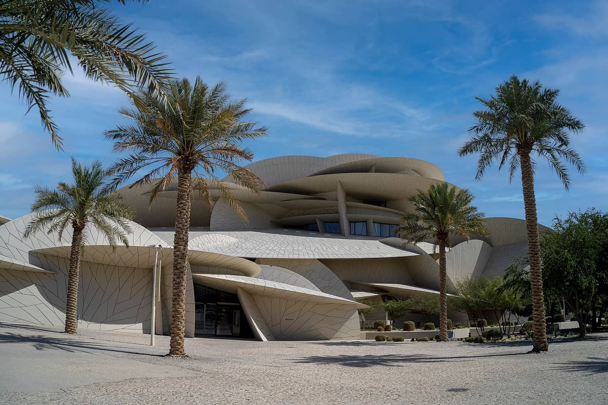 Weitwinkelansicht des Nationalmuseums von Katar mit seiner einzigartigen, von der Wüstenrose inspirierten Architektur und Palmen unter blauem Himmel.