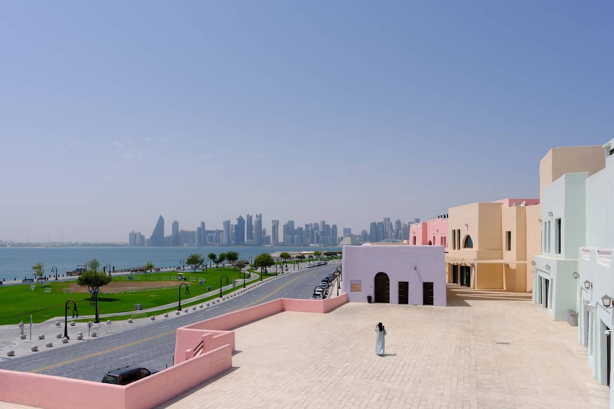 Panoramablick auf Doha mit pastellfarbenen Gebäuden, der Corniche, einem grünen Park und der Skyline von West Bay im Hintergrund unter klarem Himmel.