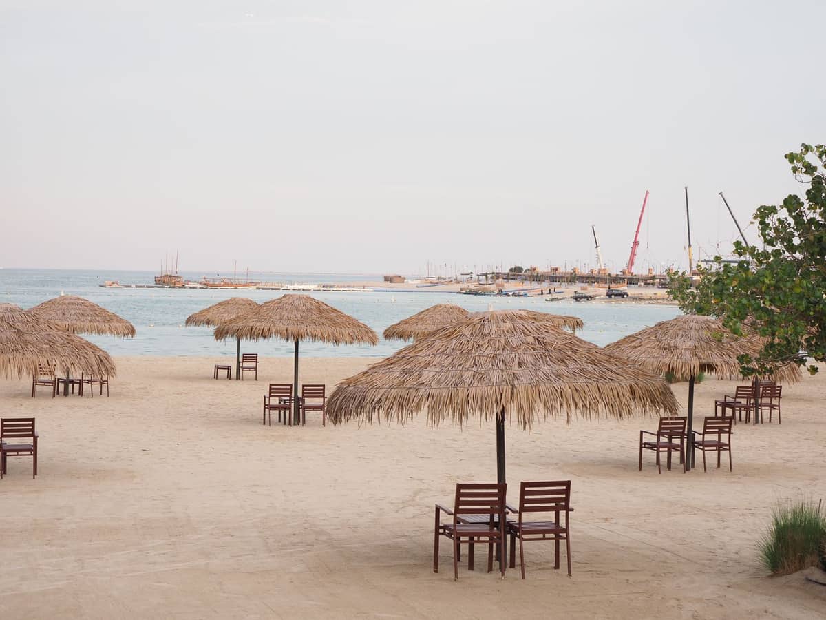 Sandstrand mit Reihen von Strohschirmen und Stühlen am ruhigen Meer, mit Booten und Kränen in der Ferne unter einem hellen Himmel.