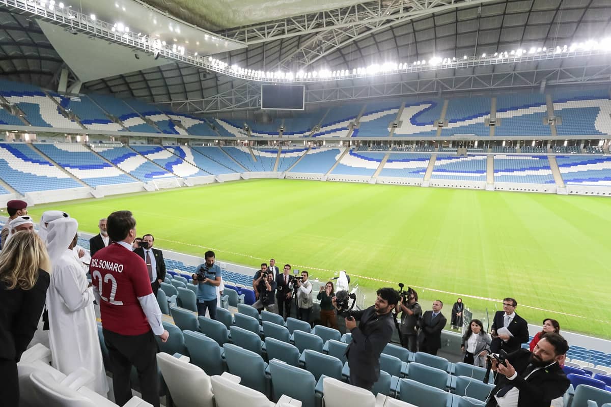 Innenansicht eines modernen Fußballstadions in Doha mit blau-weißen Sitzen und einem hellgrünen Rasen, Besucher besichtigen die Tribünen.