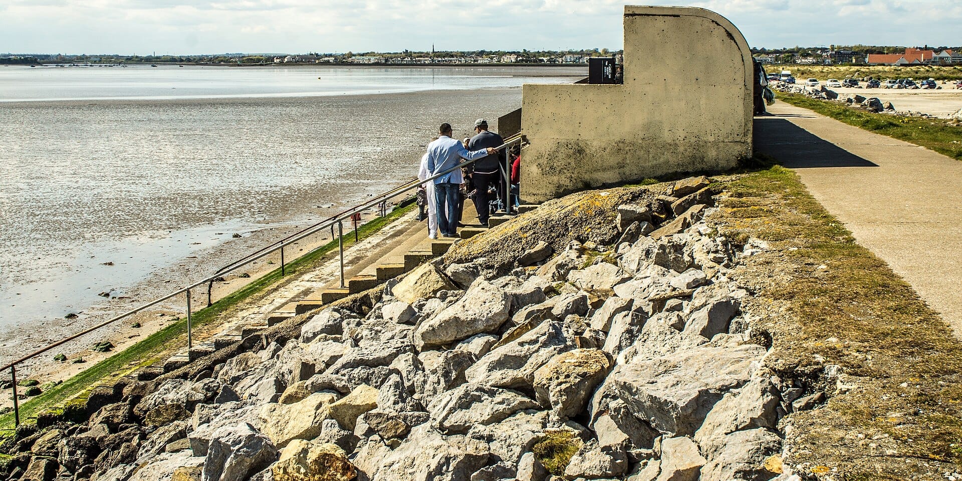 I visitatori scendono gradini in cemento dal parcheggio verso Dollymount Strand, con le distese di marea di Bull Island e le case di Dublino sullo sfondo.