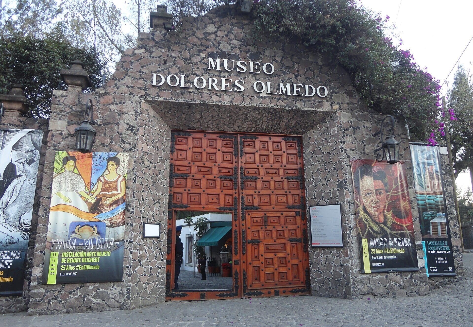 Ingresso in pietra del Museo Dolores Olmedo a Città del Messico, con un grande portone in legno, striscioni colorati e rigogliosa vegetazione sopra il cancello.