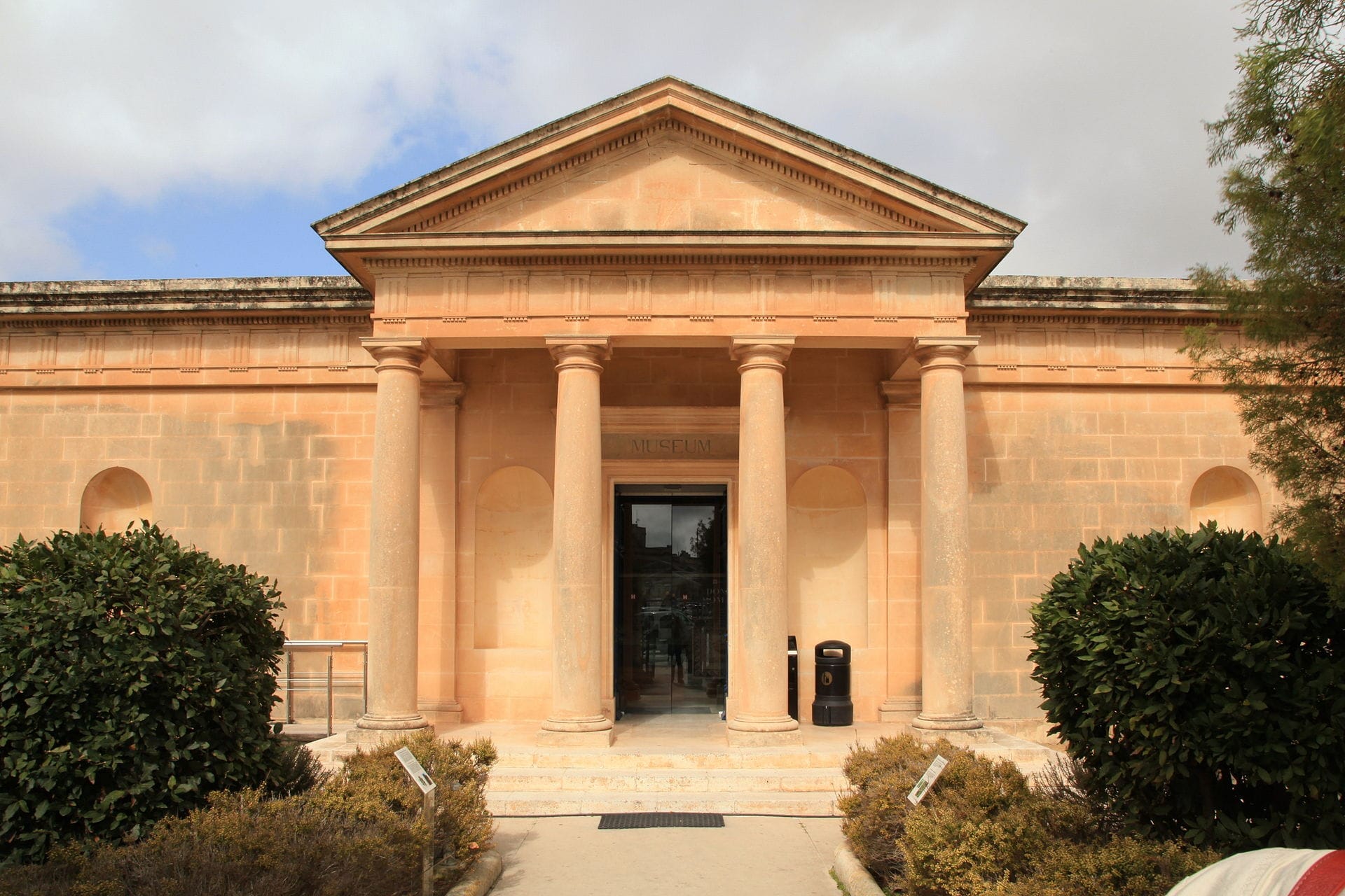 Vista frontale del museo Domus Romana a Malta, con colonne neoclassiche, pietra color sabbia e cespugli curati sotto un cielo parzialmente nuvoloso.