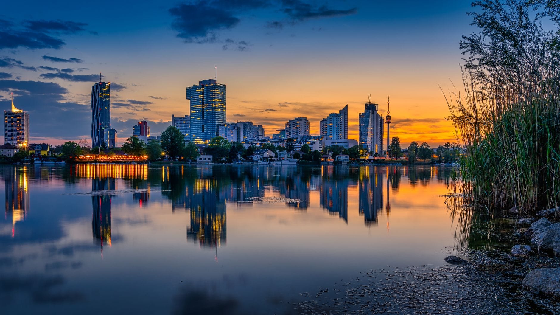 Beeindruckender Sonnenuntergang über der Donauinsel und Donaucity – moderne Hochhäuser spiegeln sich im ruhigen Donauwasser, im Vordergrund Schilf.