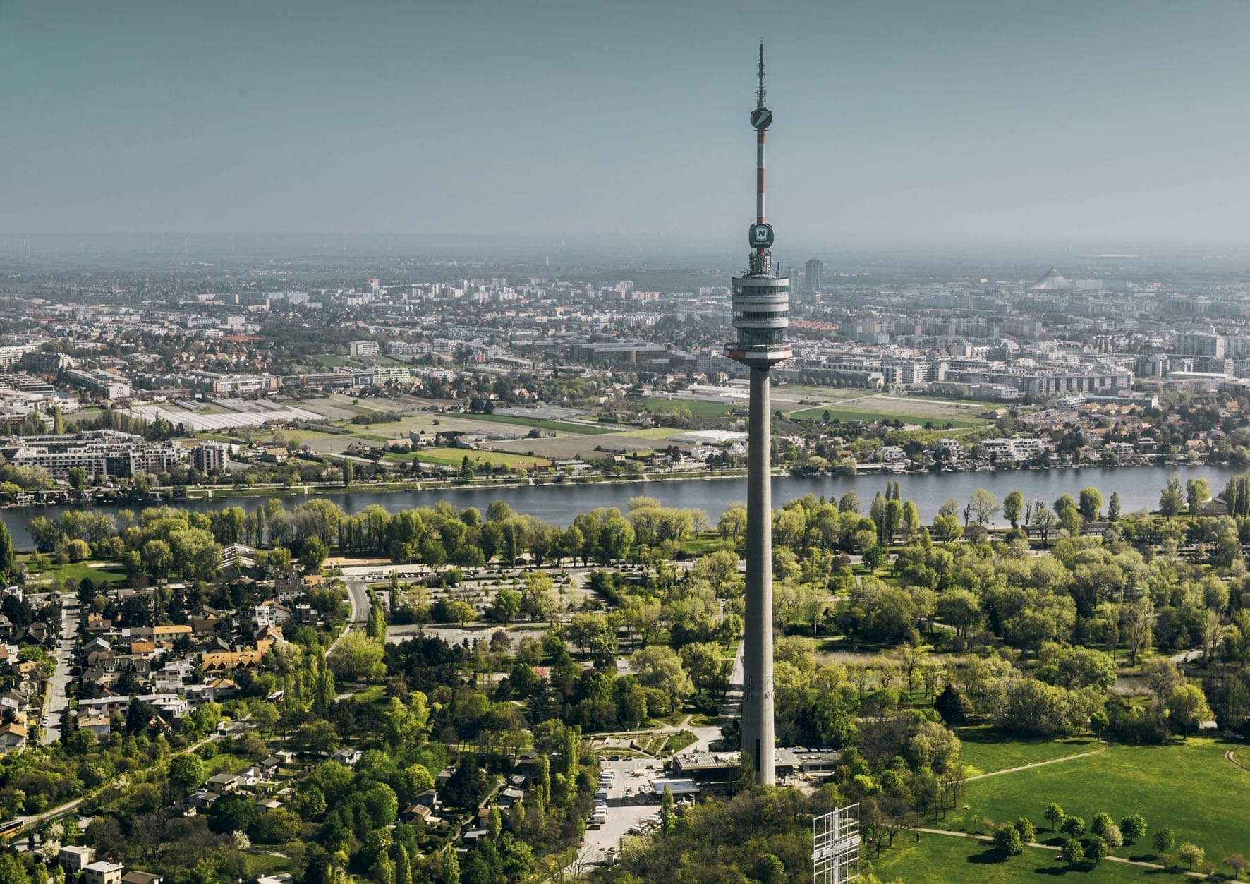 Luftaufnahme des Donauturms, der über eine üppige Parklandschaft ragt, mit der Donau und der Wiener Stadtsilhouette in der Ferne.