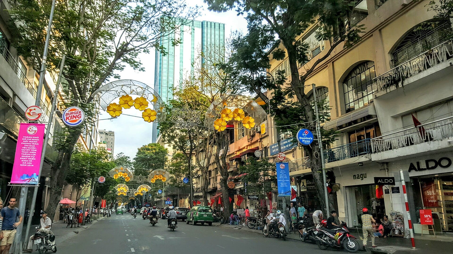 Vista de la calle Dong Khoi en Saigón, flanqueada por edificios coloniales, torres de gran altura, tiendas, luces decorativas amarillas y personas caminando.