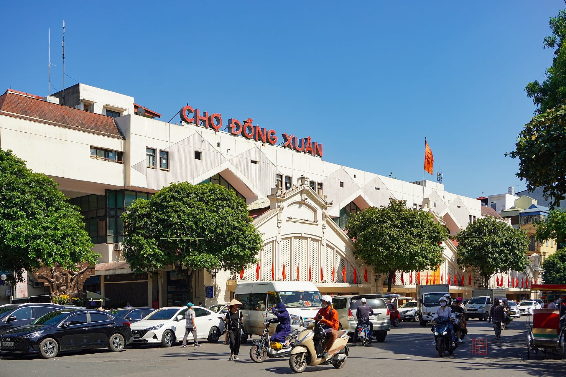 Exterior view of Đồng Xuân Market in Hanoi, featuring its prominent white facade, red signage, and bustling street traffic under a clear blue sky.