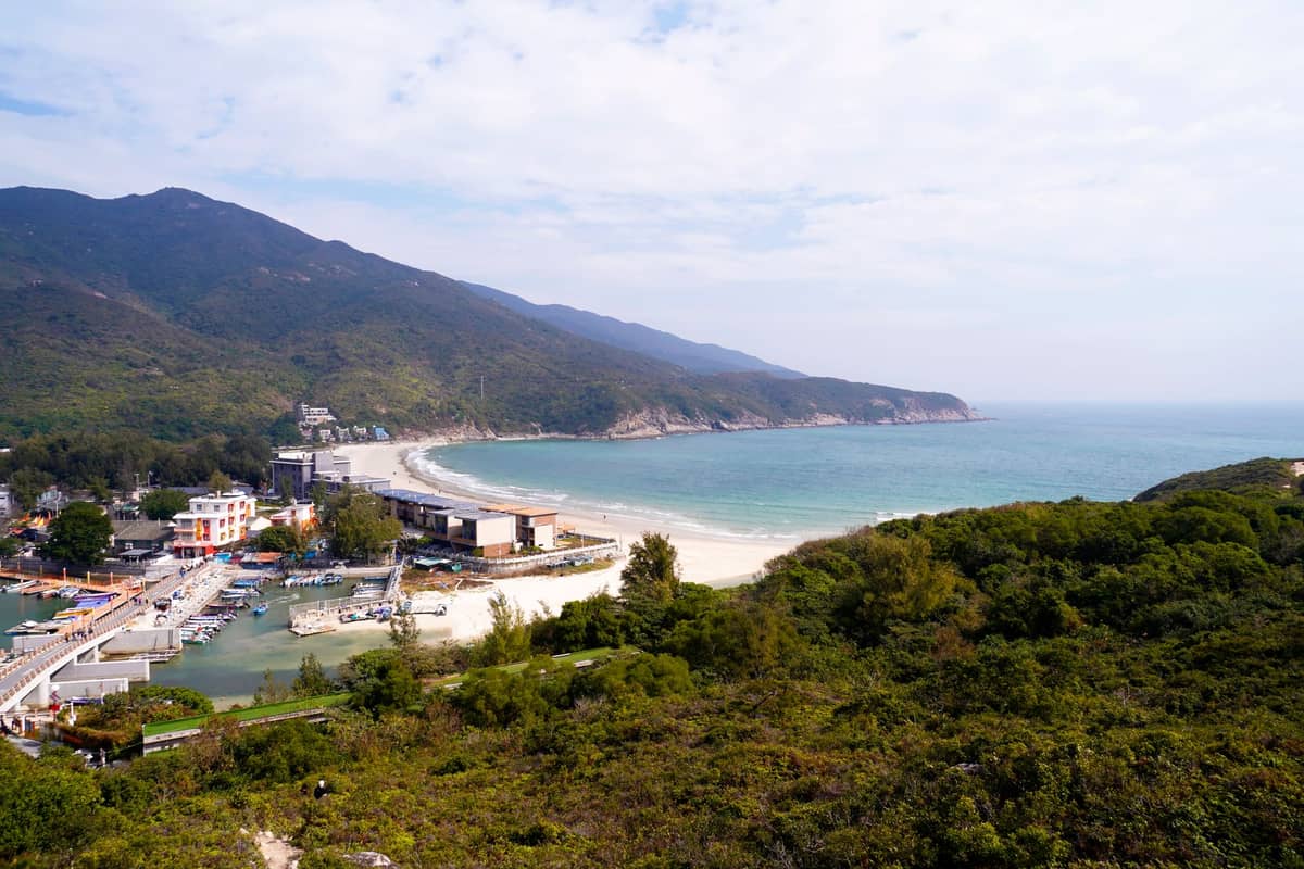 Vista panorâmica da Praia de Dongchong com areia grossa, morros imponentes, água cristalina e uma pequena vila à beira-mar no litoral leste de Shenzhen.