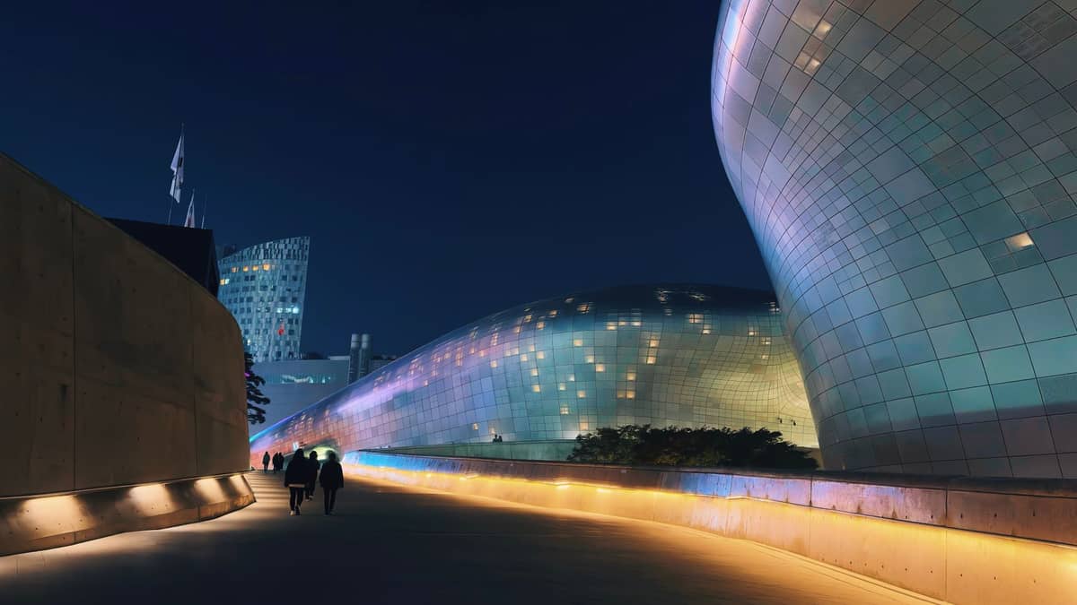 Vista noturna da arquitetura futurista e curvilínea em metal do Dongdaemun Design Plaza, com caminhos iluminados e duas pessoas caminhando pelo exterior.