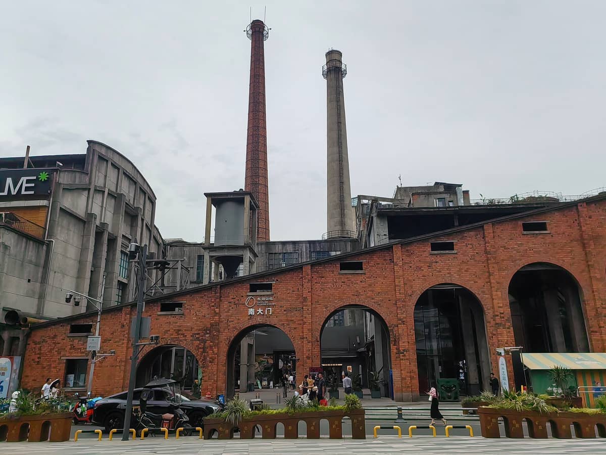 Industriegebäude aus Backstein und hohe Schornsteine im Dongjiao Memory in Chengdu, mit Menschen, die unter bewölktem Himmel spazieren.