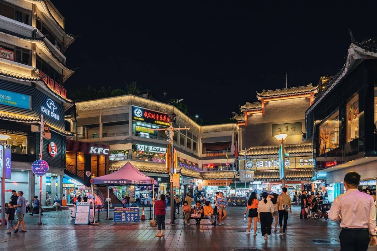 Vista noturna da Rua Pedestre Dongmen em Shenzhen, com pessoas caminhando entre edifícios tradicionais e modernos intensamente iluminados.