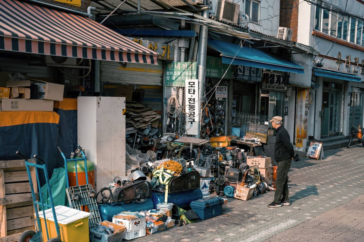 Cena de rua na Feira de Pulgas de Dongmyo, em Seul, com um senhor idoso examinando pilhas de ferramentas e objetos usados em frente a lojas tradicionais.