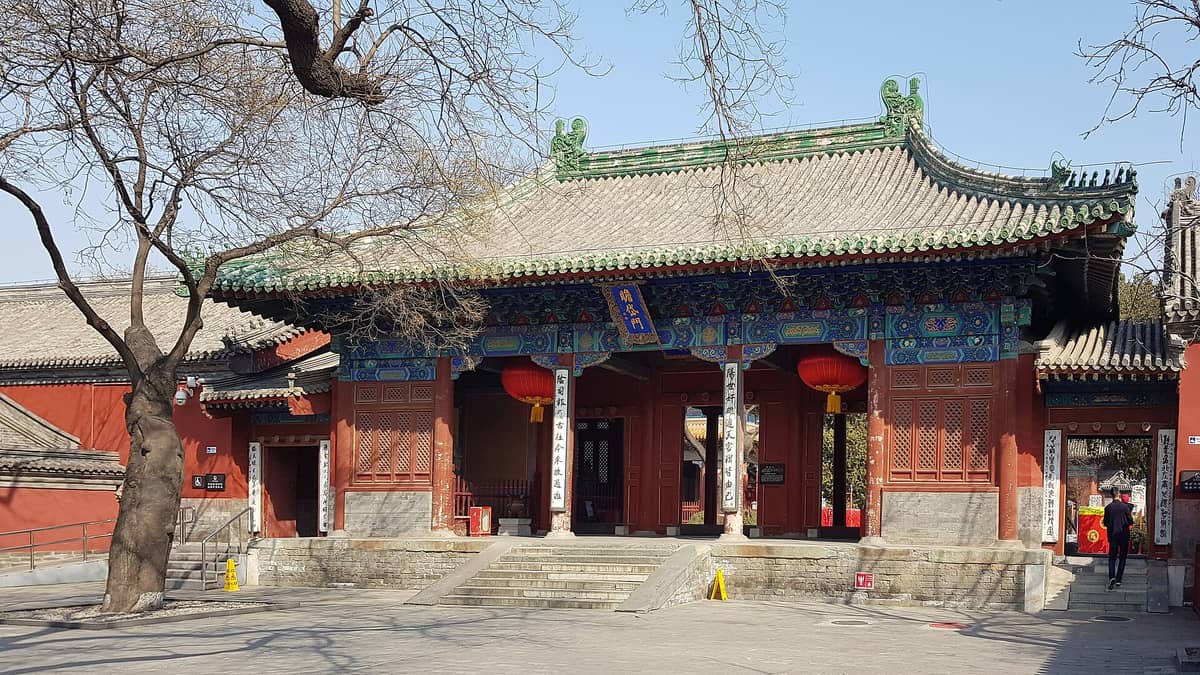 Eingangstor des Dongyue-Tempels in Peking mit traditionellen grünen Dachziegeln, buntem bemaltem Holzwerk, roten Säulen und chinesischen Laternen unter kahlen Bäumen.
