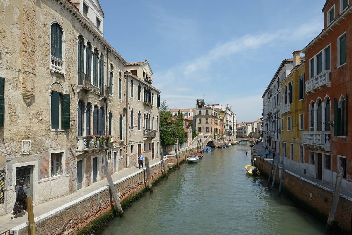 Breiter Kanal in Dorsoduro, Venedig, gesäumt von historischen, bunten Häusern und einer steinernen Fußgängerbrücke in der Ferne unter blauem Himmel.