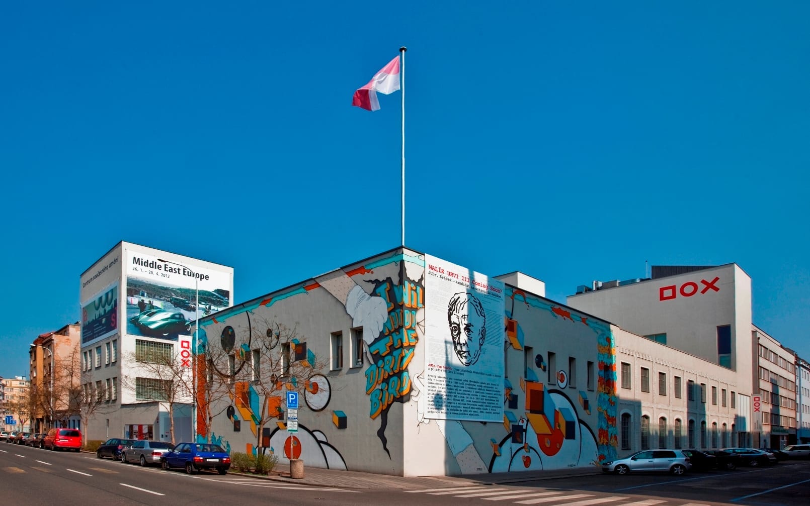 Außenansicht des DOX Centre for Contemporary Art in Prag, mit auffälligen Wandmalereien und tschechischer Flagge unter einem klaren blauen Himmel.