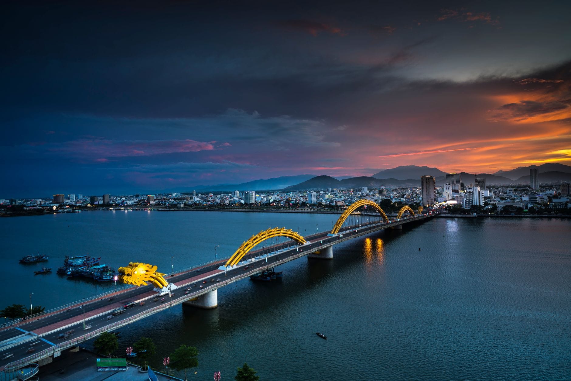 ภาพกว้างของสะพานมังกรดานังทอดข้ามแม่น้ำฮ่านช่วงพระอาทิตย์ตก ตัวมังกรสีทองเด่นเป็นสง่าพร้อมไฟส่อง สกายไลน์เมืองฉากหลัง