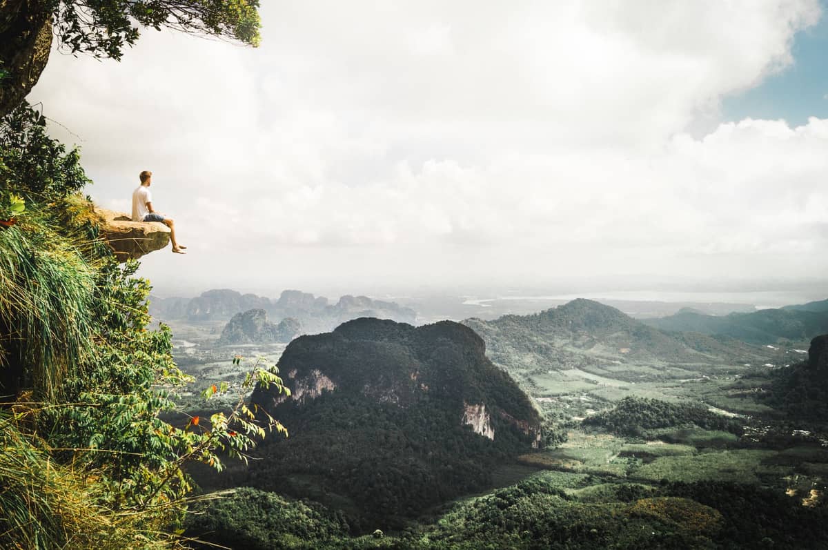 Eine Person sitzt auf dem felsigen Rand des Dragon Crest Mountain und blickt auf ein saftig grünes Tal und Kalksteinfelsen mit einem weiten Panoramablick in Krabi, Thailand.