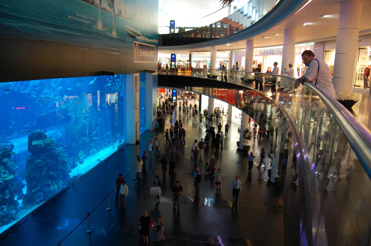 Weitwinkelaufnahme des riesigen Glasbeckens des Dubai Aquariums im Dubai Mall, mit Besuchern im Mall-Bereich und dem mit Fischen gefüllten Aquarium.