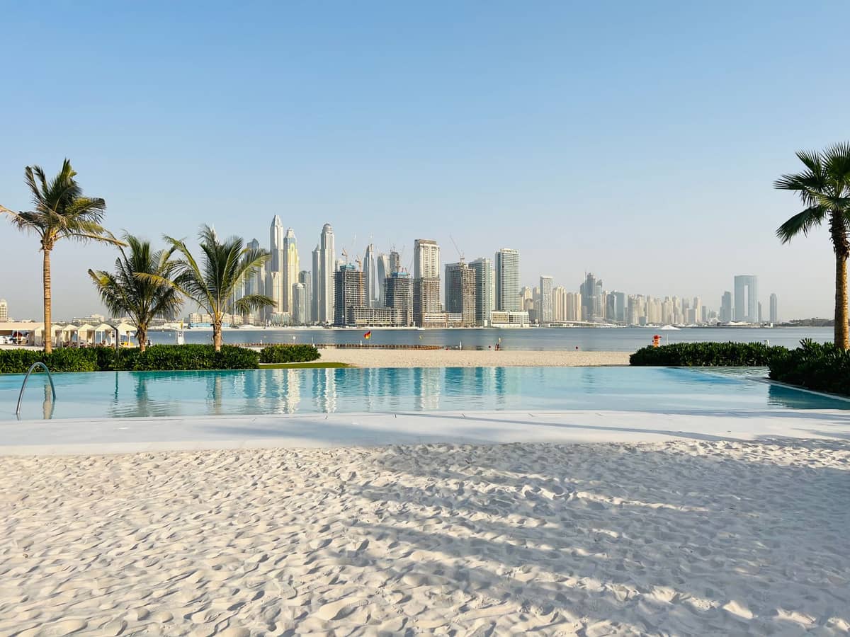 Ein luxuriöser Dubai Beach Club mit Pool, weißem Sand und Palmen – mit Blick auf die ikonische Dubai Marina Skyline an einem sonnigen, klaren Tag.