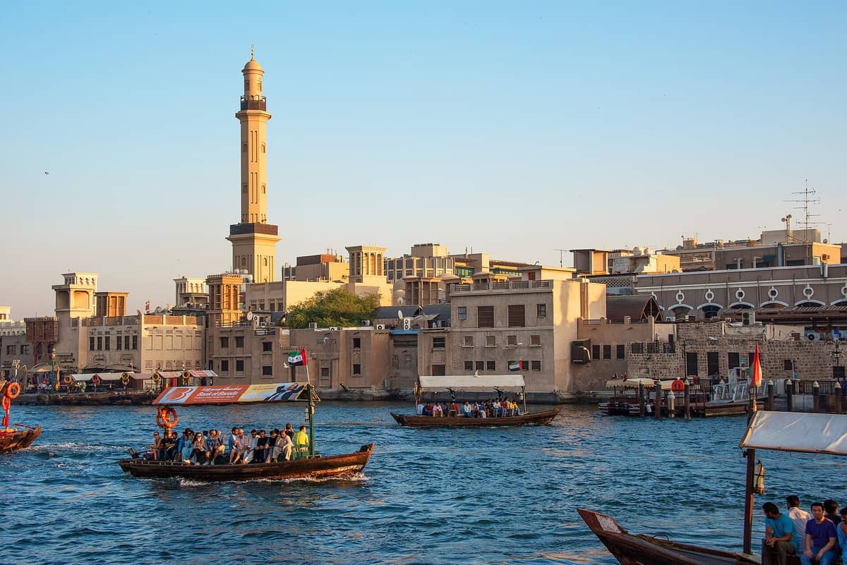 Traditionelle Abras mit Passagieren auf dem Dubai Creek, im Hintergrund historische Gebäude und das markante Minarett einer Moschee unter strahlend blauem Himmel.
