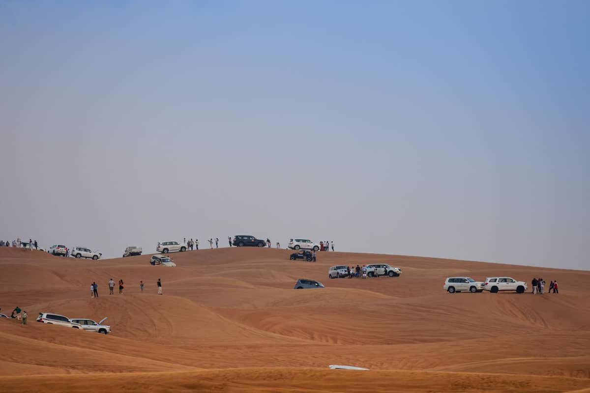 Konvoi weißer 4x4-Fahrzeuge, die über Sanddünen fahren und anhalten, mit Gruppen von Menschen, typisch für ein Wüstensafari-Erlebnis in Dubai.