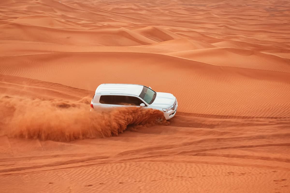 Ein weißer SUV wirbelt orangefarbenen Wüstensand auf, während er bei einer Dubai-Wüstensafari über hohe Dünen fährt.