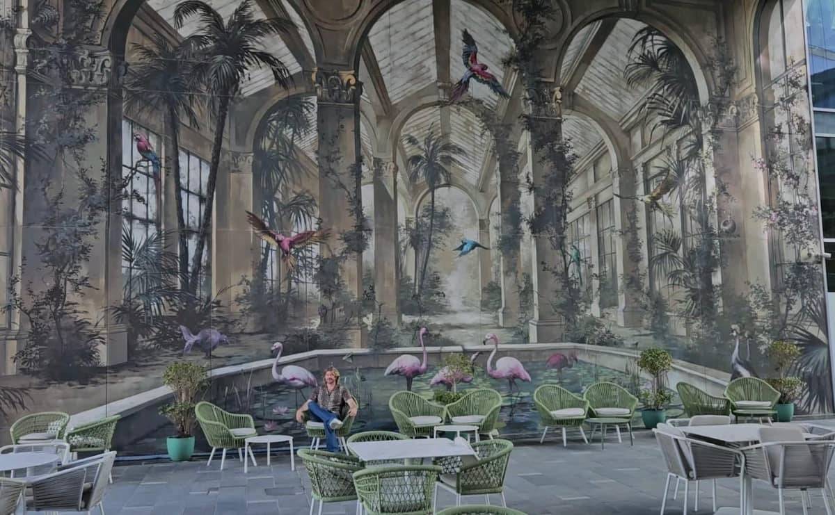 Outdoor-Cafébereich im Dubai Design District mit einem großen Wandgemälde, das tropische Vögel und Flamingos in einem kunstvollen Gewächshaus zeigt, umgeben von grünen Stühlen und Tischen.
