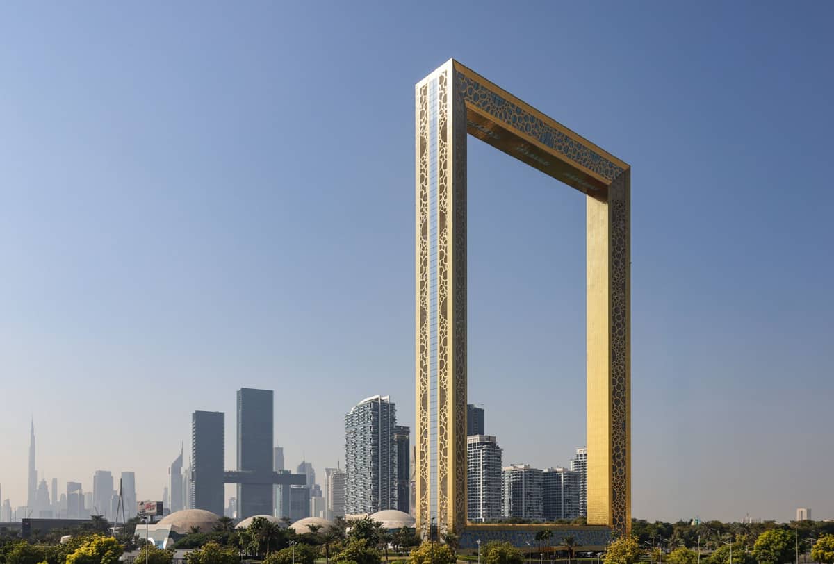 Weitwinkelaufnahme des Dubai Frame, einer riesigen goldenen rechteckigen Struktur im Zabeel Park, mit der modernen Stadtsilhouette im Hintergrund unter einem strahlend blauen Himmel.