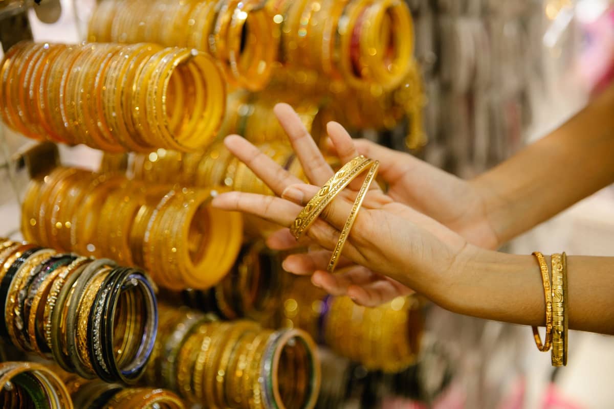 Person, die in einem Geschäft goldene Armreife anprobiert, hinter ihr eine Auslage voller Goldschmuck.