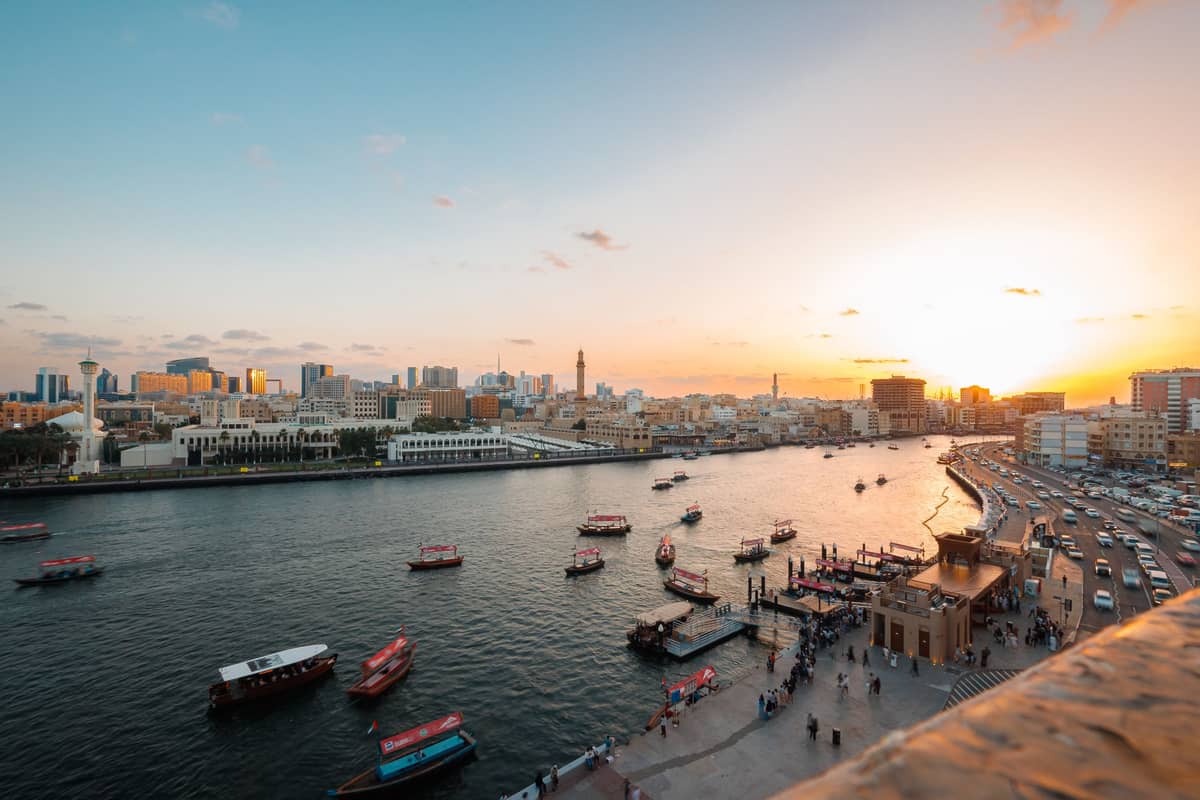 Weitläufiger Blick über den Dubai Creek bei Sonnenuntergang mit traditionellen Booten, historischen Gebäuden und einer Uferpromenade, der das ruhige lokale Leben abseits der Massen zeigt.