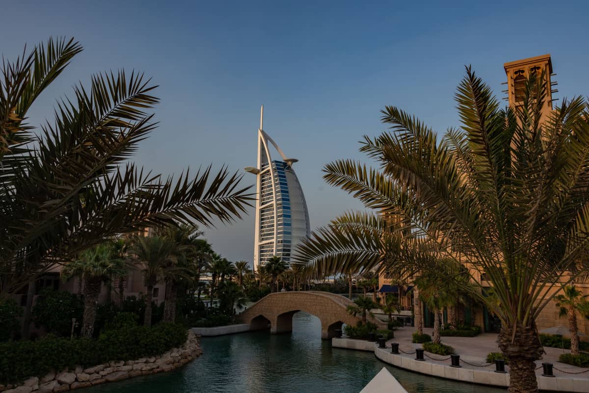Blick auf das Hotel Burj Al Arab, eingerahmt von Palmen und einem Wasserweg, mit einer Steinbrücke im Vordergrund bei Sonnenuntergang.
