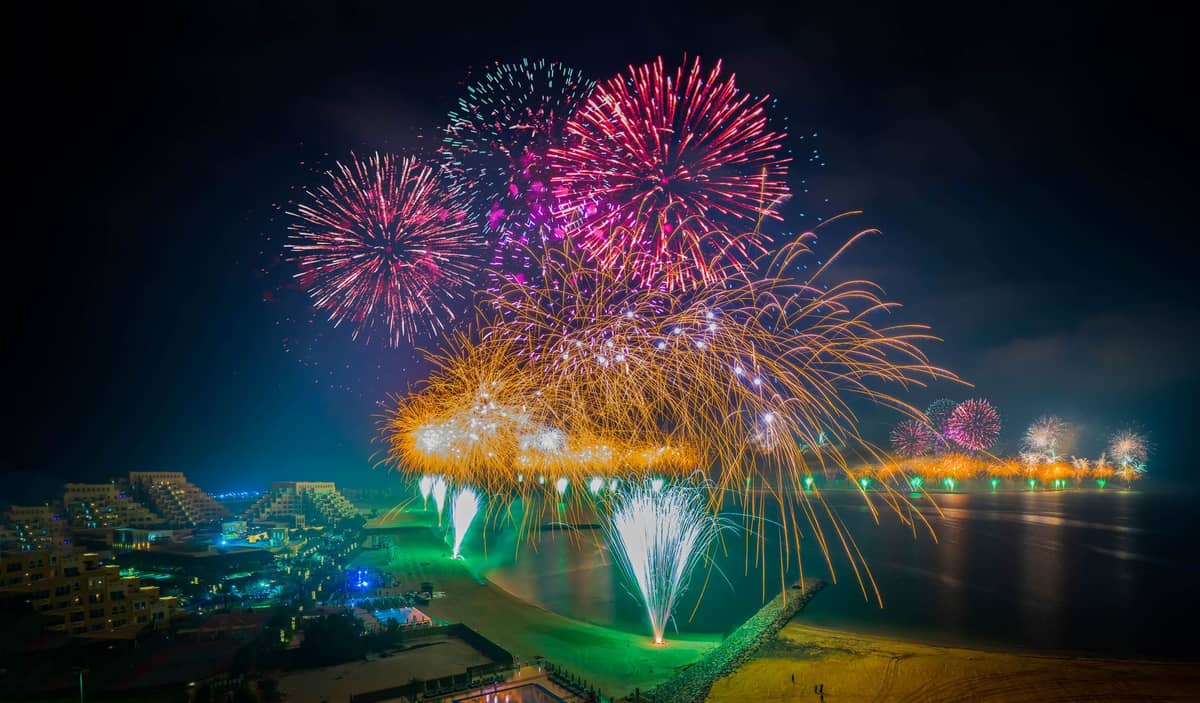 Bunte Feuerwerke über einer Reihe von Strandhotels und Resorts in Dubai bei Nacht, mit Spiegelungen auf dem Wasser.