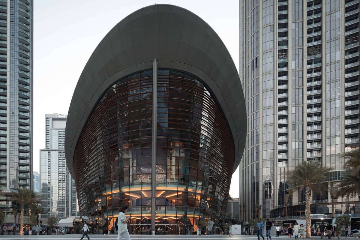 Frontansicht der Dubai Opera – ein modernes Glas-und-Stahl-Gebäude in Form eines traditionellen Daus, umgeben von Wolkenkratzern in Downtown Dubai, mit Menschen auf der Plaza davor.