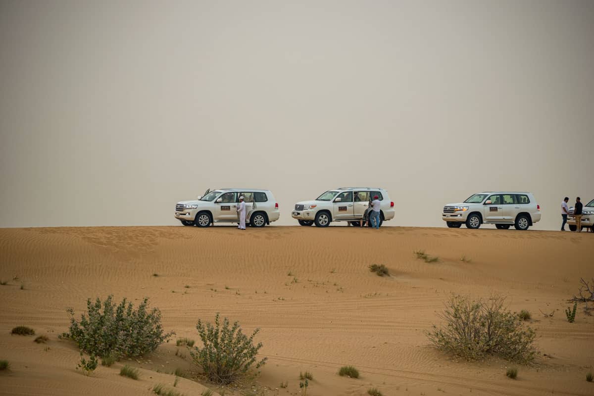 Drei weiße SUVs auf goldenen Sanddünen in der Wüste von Dubai geparkt, mit Menschen in der Nähe unter einem diesigen Himmel.