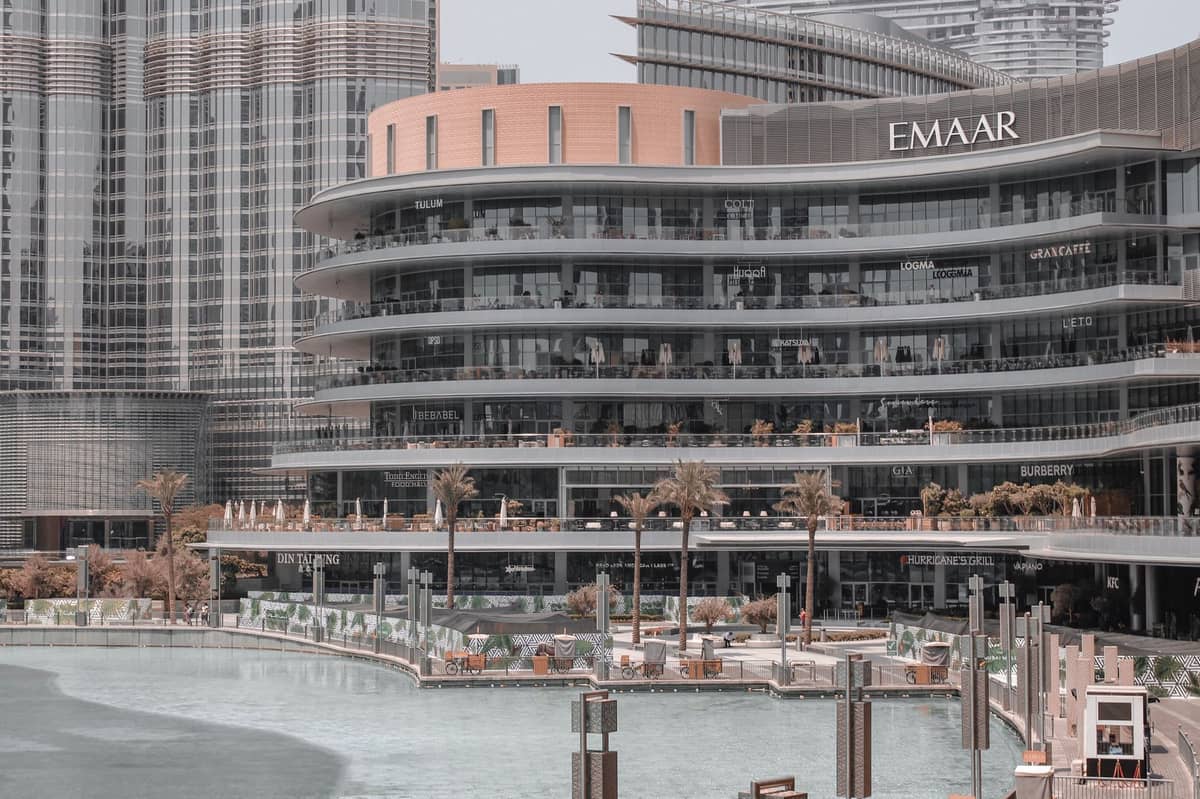 ศูนย์การค้าสมัยใหม่ในดูไบที่มีป้าย EMAAR ระเบียงกลางแจ้ง และผู้คนบริเวณริมน้ำ ล้อมรอบด้วยตึกระฟ้าสูงใหญ่