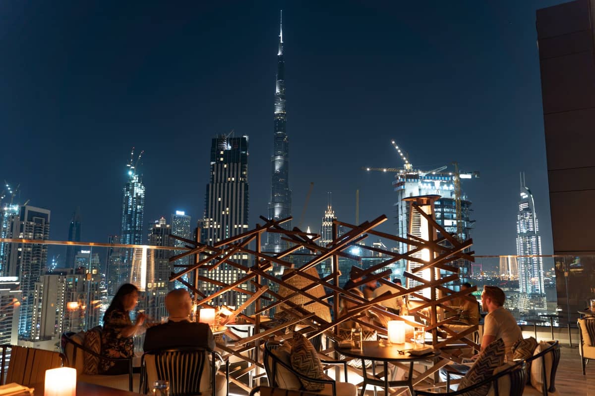 Nächtlicher Blick von einer Rooftop-Bar in Dubai mit Gästen beim Essen, dem beleuchteten Burj Khalifa und der Skyline im Hintergrund.