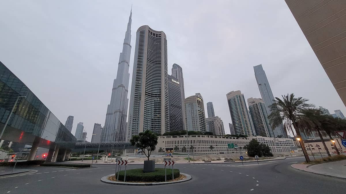 Weitwinklige Straßenansicht des Downtown Dubai mit dem Burj Khalifa, Metro-Gehweg, Wolkenkratzern, Palmen und städtischen Straßenschildern unter bewölktem Himmel.