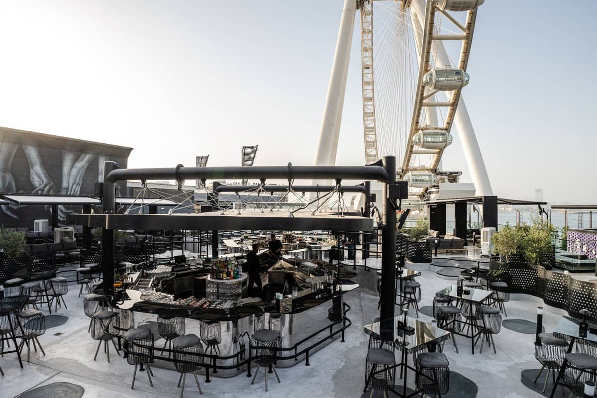 Modernes Dachbar in Dubai mit Tischen und zentralem Barbereich, in der Nähe eines großen Riesenrads gelegen, bei hellem Tageslicht fotografiert.