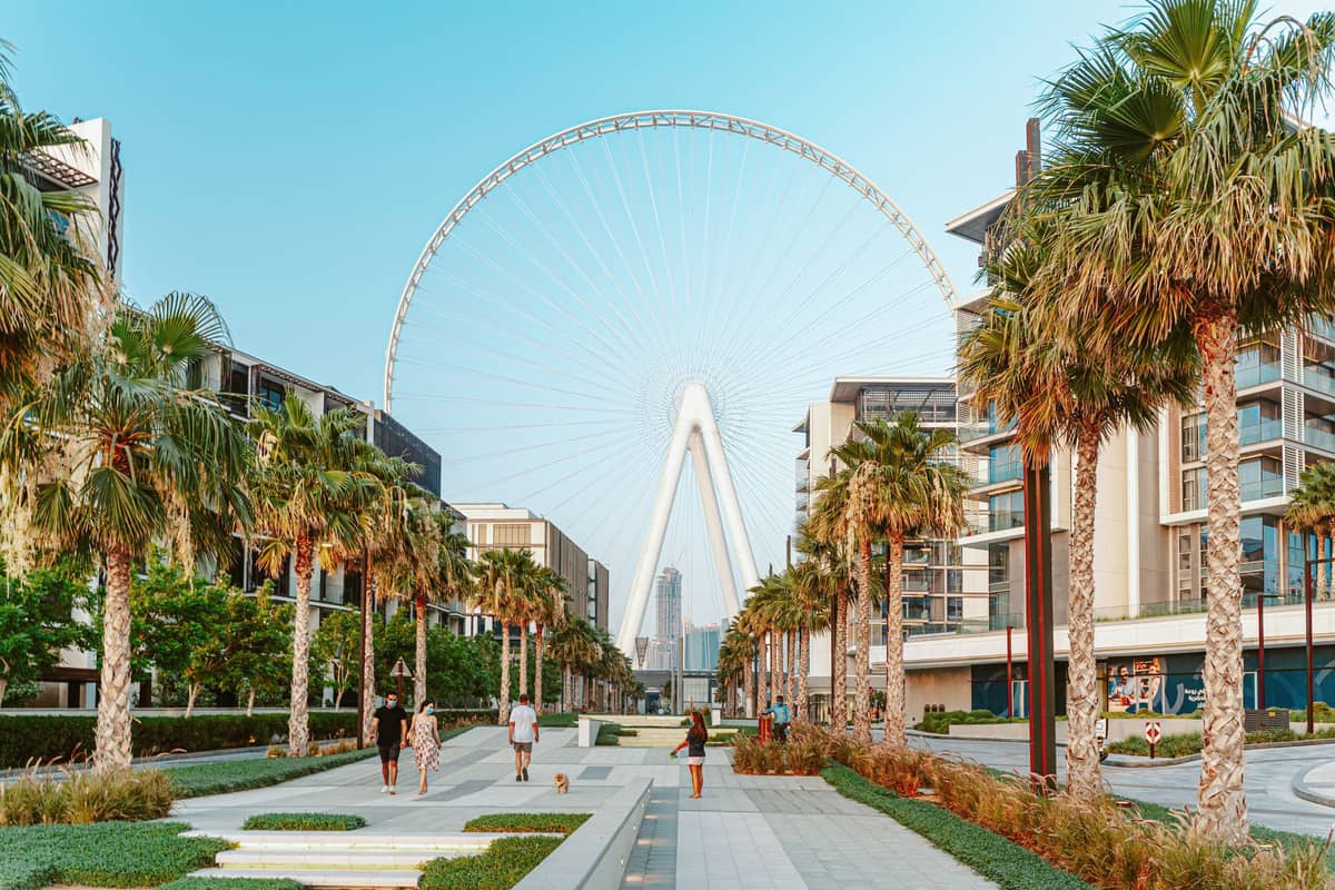 Menschen schlendern entlang einer modernen Promenade in Dubai, gesäumt von Palmen, mit dem Riesenrad Ain Dubai im Hintergrund an einem klaren Tag.