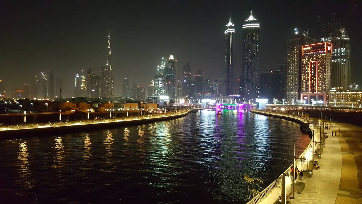 Nachtansicht des Dubai Water Canal mit beleuchteter Promenade, Skyline im Hintergrund und bunten Lichtreflexen auf dem Wasser.