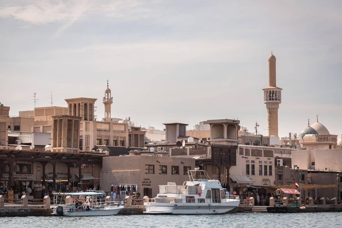 Blick über den Dubai Creek mit traditionellen Windturm-Gebäuden, Minaretten, Booten und lebhaftem Treiben an der Uferpromenade unter einem strahlend blauen Himmel.