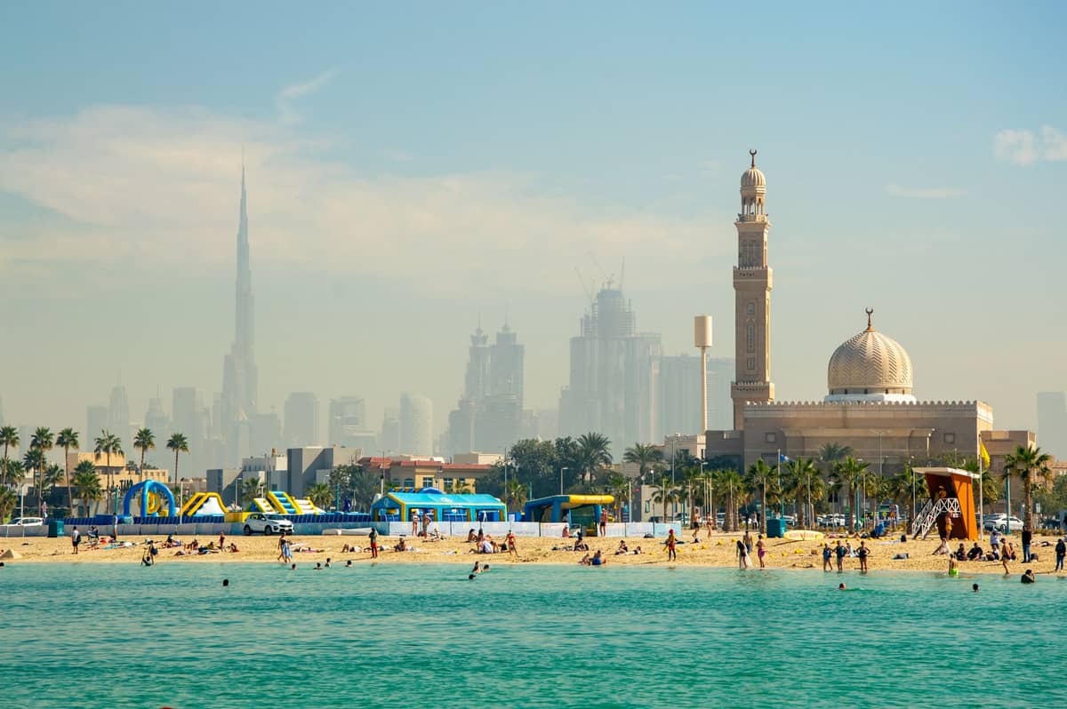 Familien und Kinder genießen einen Sandstrand in Dubai mit Wasseraktivitäten, einer Moschee, Palmen und der Skyline von Dubai im Hintergrund.