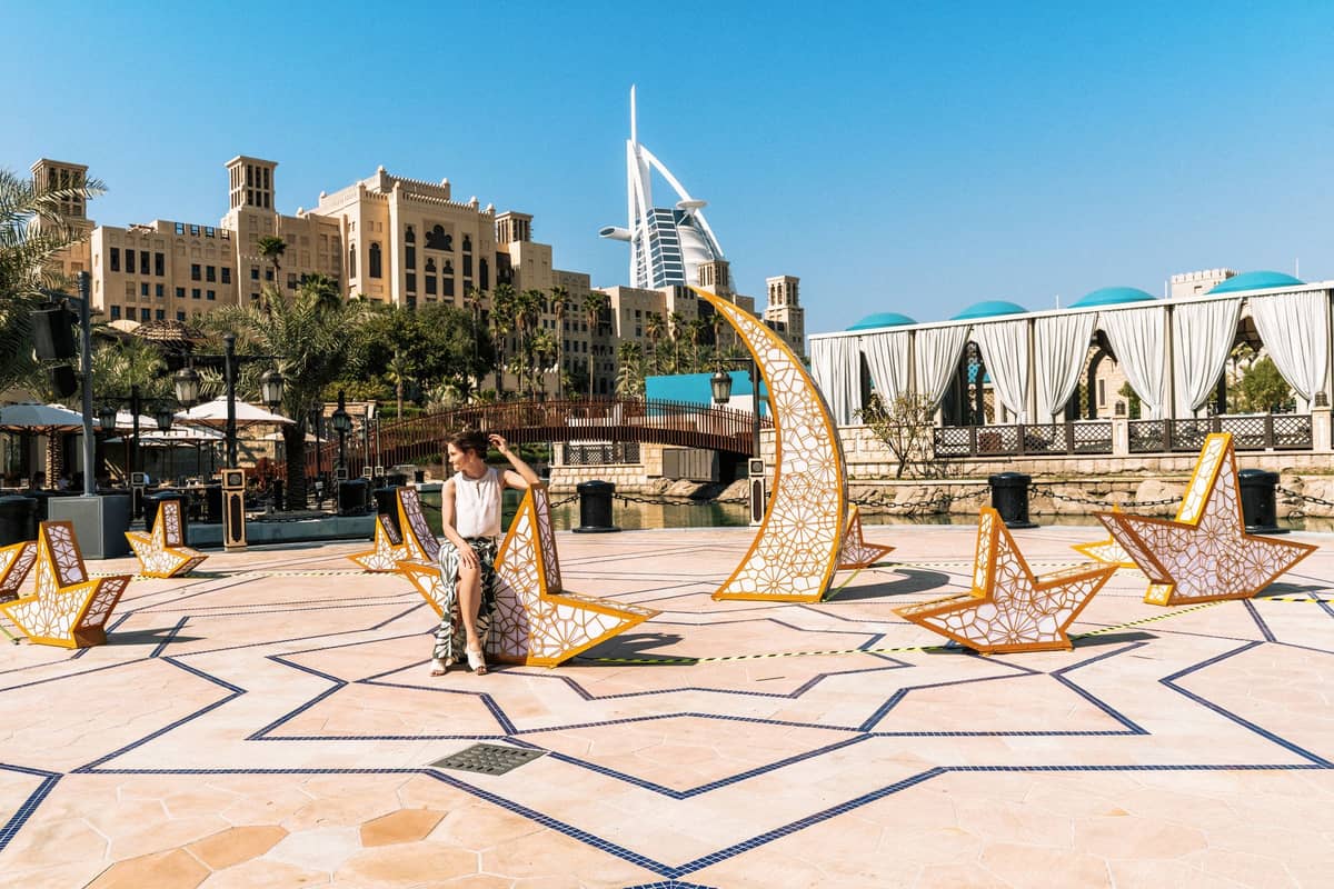 Außenplatz in Dubai mit dekorativen Stern- und Halbmondskulpturen, sitzendes Kind, Burj Al Arab und traditionelle Gebäude im Hintergrund