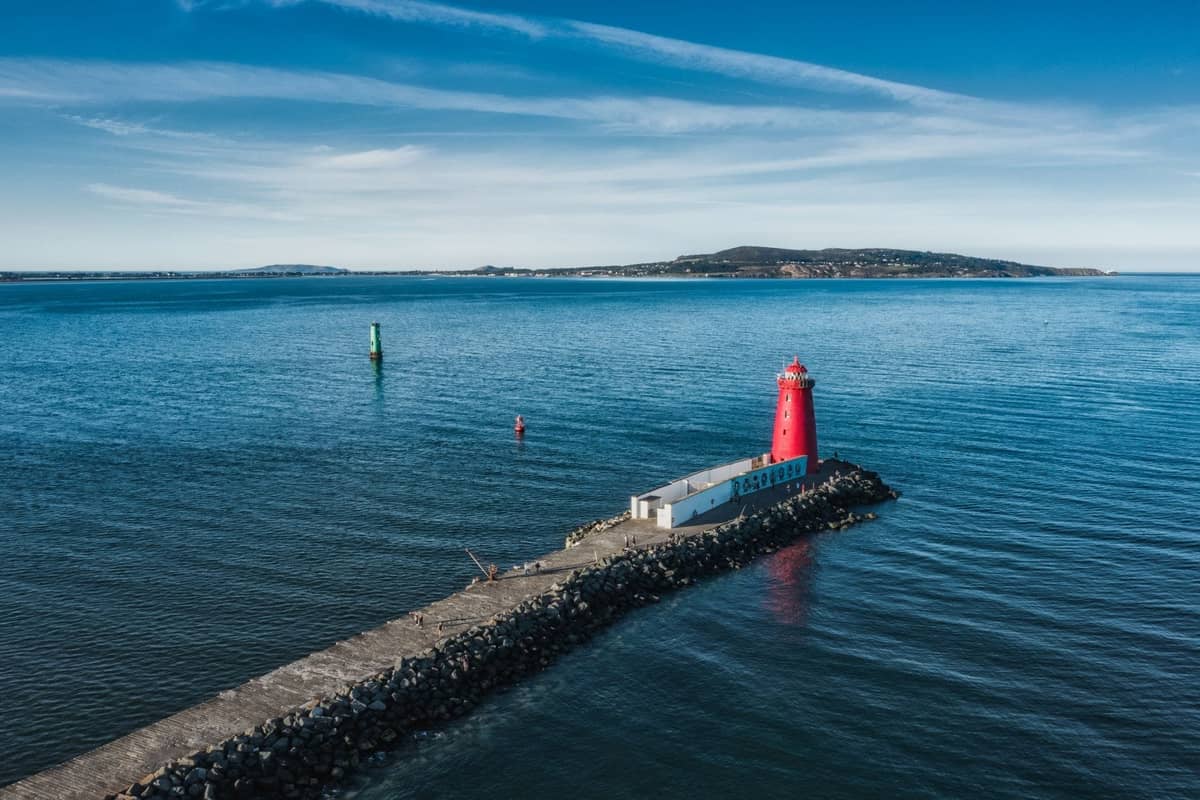 Luftaufnahme der Great South Wall in der Dublin Bay, die am ikonischen roten Poolbeg Lighthouse endet, mit blauem Meer und Land in der Ferne.