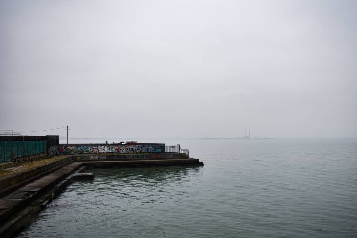 Ein grauer, bewölkter Tag an einem Küstenpier in Dublin, mit einer Graffitiwand und ruhiger See. Die Poolbeg-Schornsteine sind am Horizont schwach zu erkennen.