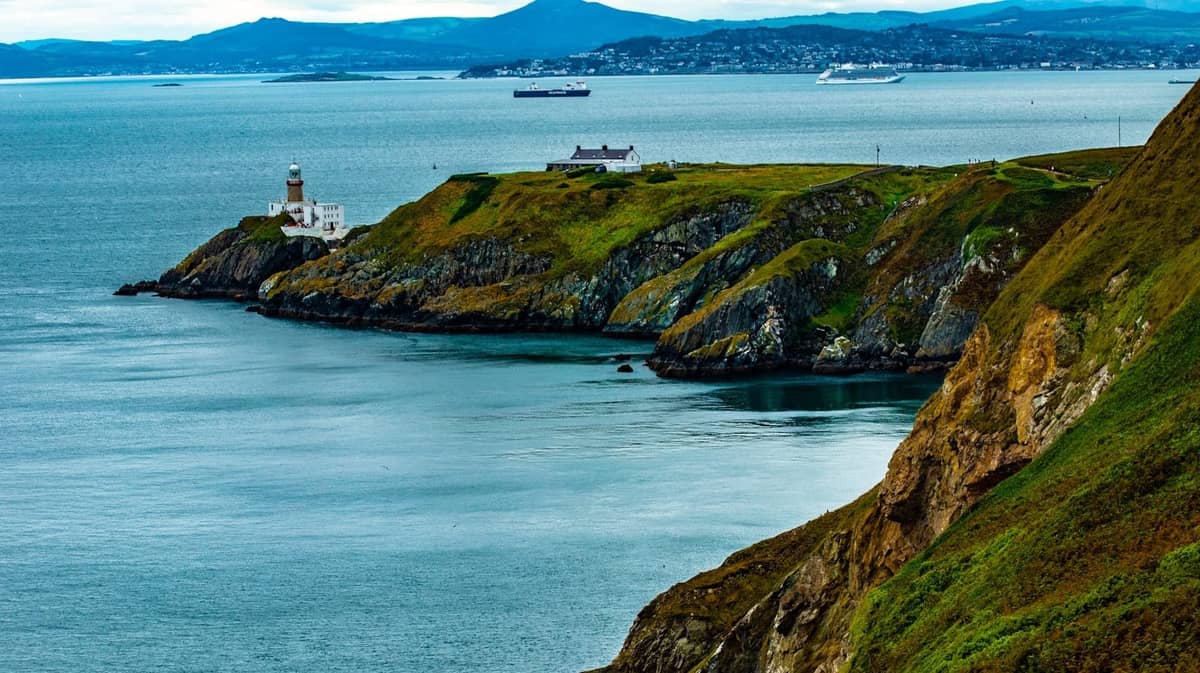 Malerische Küstenklippen von Howth mit einem Leuchtturm mit Blick auf die Dublin Bay und entfernte Hügel unter einem teilweise bewölkten Himmel.