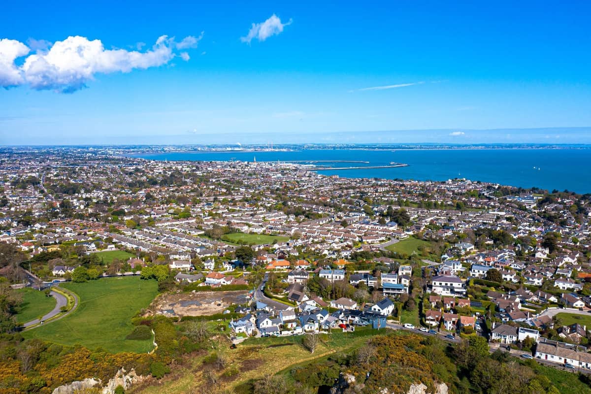 Weite Luftaufnahme eines Küstendorfes in der Nähe von Dublin mit Häusern, Grünflächen und dem Meer, das sich unter einem blauen Himmel in die Ferne erstreckt.