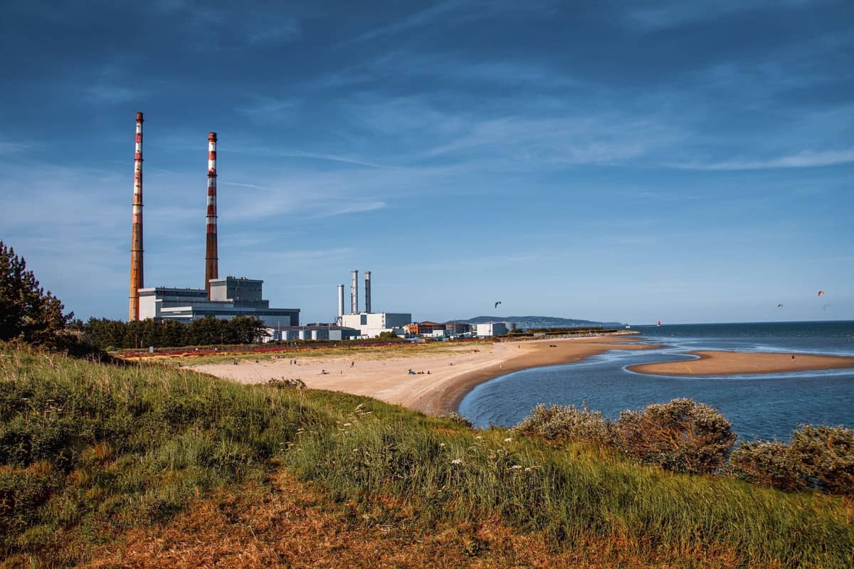 Breiter Sandstrand nahe Dublin mit grasbewachsenen Dünen, geschwungener Küstenlinie und den Poolbeg-Schornsteinen im Hintergrund unter blauem Himmel.