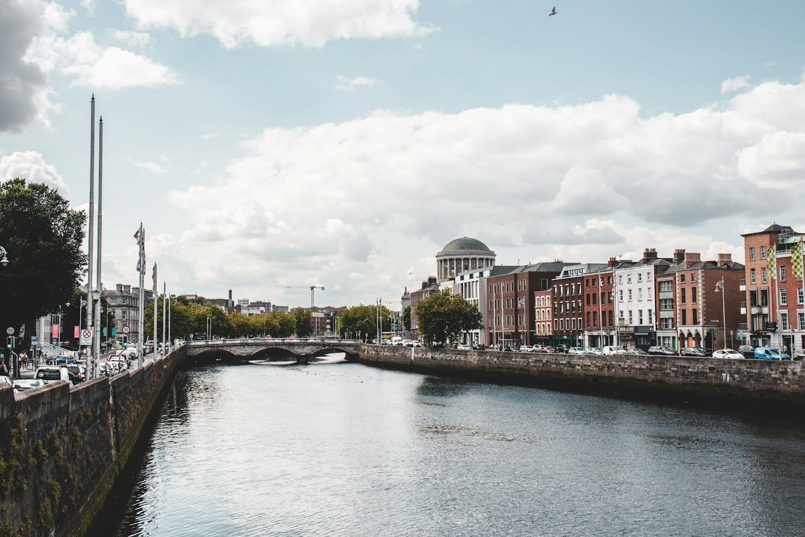 Vista diurna do Rio Liffey em Dublin com prédios georgianos clássicos, uma ponte de pedra e a cúpula dos Four Courts sob um céu parcialmente nublado.